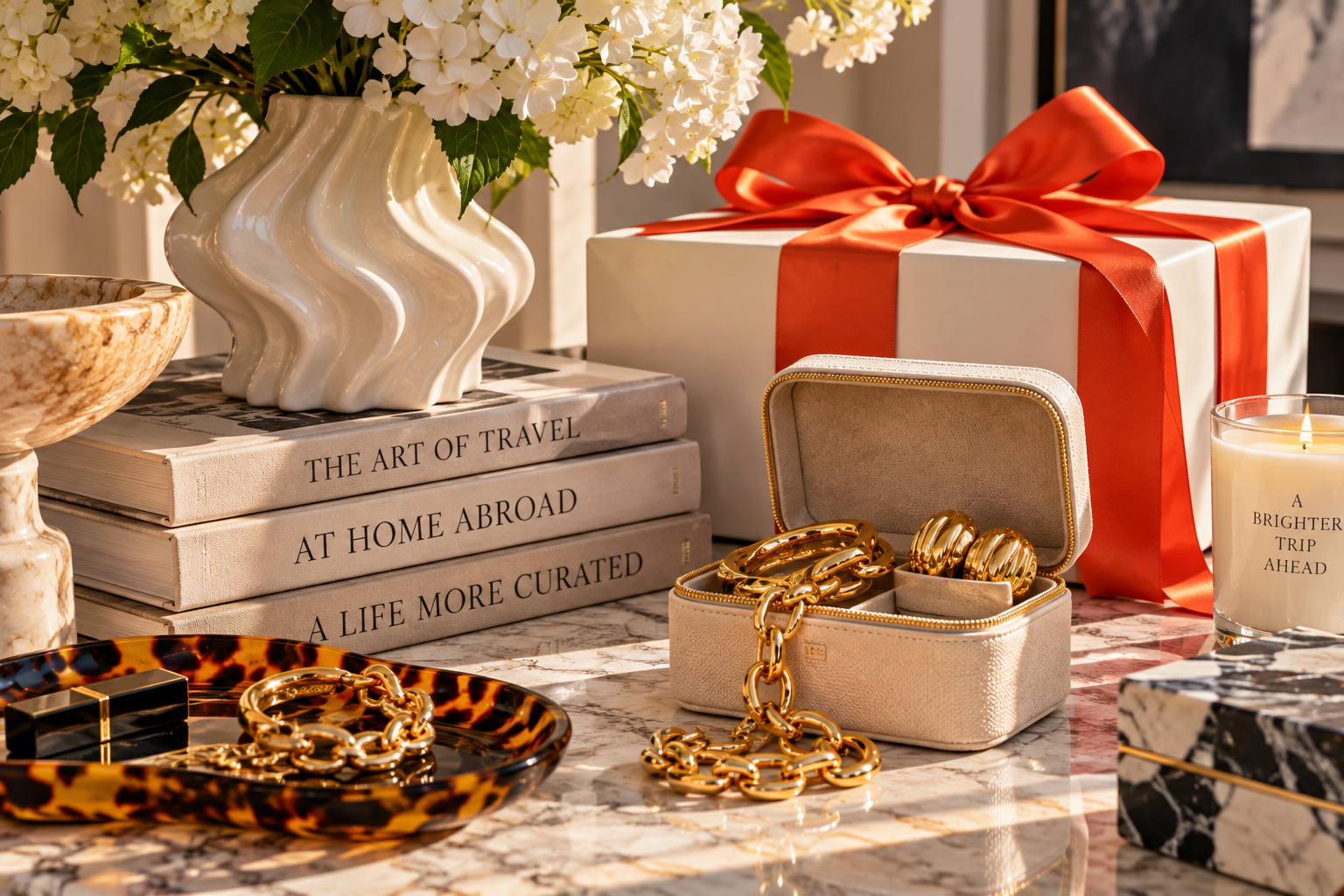 A luxury gift still life with white flowers, neutral books, a cream box tied in red-orange ribbon, an open jewellery case, chunky gold jewellery, a tortoiseshell tray and a candle